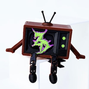 May include: A brown and black 3D-printed retro television figure with arms and legs. The TV screen displays a green and purple graphic design. The figure has two antennas and two green circular buttons.