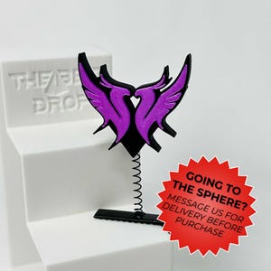 May include: A black and purple decorative piece featuring a stylized bird design with outstretched wings. The design is mounted on a spring and a black base. A red starburst-shaped sticker with text is visible.