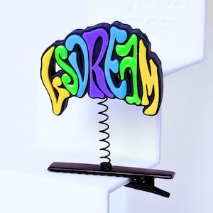 Peut inclure: Une pince à cheveux composée d'une base en métal noir de type crocodile reliée par un fin ressort en spirale noir à un ornement en plastique multicolore. L'ornement affiche le mot "DREAM" dans une police bulle avec du jaune, bleu, violet et vert.