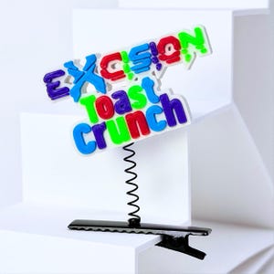 May include: A hair clip featuring a black metal alligator clip base attached to a thin black metal spring. The top of the spring holds a white plastic sign with the text EXCISION TOAST CRUNCH in raised, multi-colored letters in blue, red, and green.