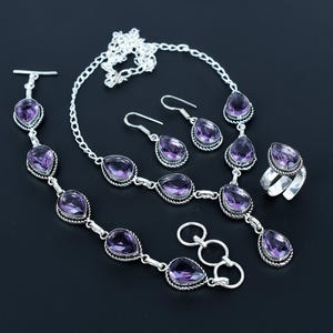Op de afbeelding: Een complete sieradenset met een ketting, armband, oorbellen en ring. Elk stuk heeft een zilverkleurige metalen zetting en is voorzien van een grote, druppelvormige, paarse edelsteen. De set wordt gepresenteerd op een zwart oppervlak.