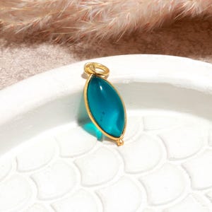 May include: A marquise-shaped pendant featuring a smooth, translucent teal stone encased in a thin gold-toned metal frame. The pendant includes a small circular gold-toned jump ring at the top for attachment to a chain or cord.