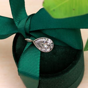 May include: A pear-shaped, clear gemstone pendant with a silver-colored bail. The pendant is set against a dark green velvet jewelry box with a matching green ribbon. A green leaf is visible in the background.