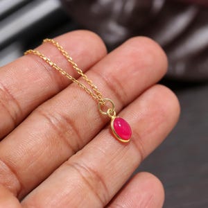 Op de afbeelding: Een goudkleurige ketting met een kleine, ovale, felroze cabochon hanger. De hanger zit vast in een eenvoudige goudkleurige metalen zetting en hangt aan een fijne, dunne schakelketting die op de huid rust.
