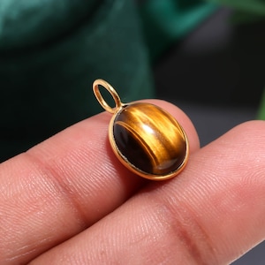 Op de afbeelding: Een gouden hanger met een ovale tijgeroogsteen. De steen heeft een goudbruine kleur met donkerbruine strepen. De hanger heeft een goudkleurige bezel setting en een kleine lus aan de bovenkant voor een ketting.