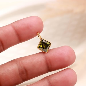 May include: A small square-shaped pendant featuring a faceted olive green gemstone set in a gold-toned metal frame. The pendant includes a small circular jump ring at the top for attachment, showcasing a clear, polished finish and geometric design.