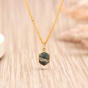 May include: A gold-toned chain necklace featuring a small, hexagonal pendant. The pendant contains a dark green and grey moss agate stone with visible natural inclusions, encased in a smooth, polished gold-coloured metal frame.