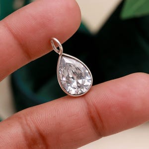 Può includere: Un ciondolo a forma di pera con una grande gemma trasparente e sfaccettata incastonata in una montatura in metallo argentato. Il ciondolo ha un piccolo anello nella parte superiore per una catena. La gemma brilla.