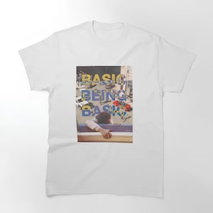 Könnte beinhalten: Weißes klassisches T-Shirt mit einem Grafikdesign. Das Design zeigt die Worte "BASIC BEING BASIC" in Gelb und Blau und eine Person, die sich auf einer Brüstung abstützt, mit einem Stadtbild im Hintergrund.