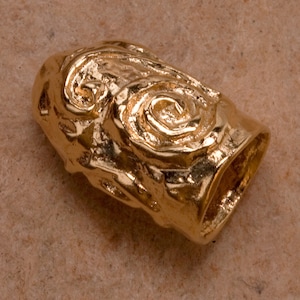 Puede incluir: Una cuenta de metal de color dorado con un diseño de remolino y textura. La cuenta tiene un agujero en el centro, lo que sugiere que está destinada a ser enhebrada en un collar o pulsera.