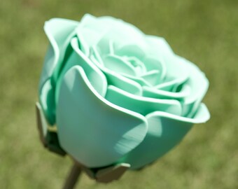 Può includere: Primo piano di una rosa verde menta con petali dettagliati. Il fiore artificiale ha uno stelo ed è ambientato su uno sfondo verde sfocato, che suggerisce un ambiente da giardino. La rosa è in piena fioritura.