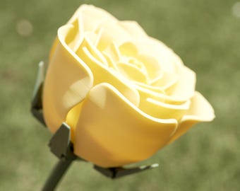 Può includere: Primo piano di una rosa gialla in piena fioritura. I petali sono di un giallo morbido e cremoso, su uno sfondo verde sfocato. Lo stelo è verde scuro, con piccole foglie verdi alla base del fiore.