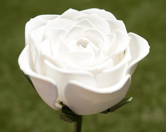 Può includere: Primo piano di una rosa bianca con numerosi petali. La rosa è in piena fioritura, con una consistenza dettagliata. Lo stelo è verde scuro e lo sfondo è verde sfocato.