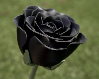 Può includere: Una rosa nera dettagliata con petali a strati e uno stelo verde. Il fiore artificiale è in piena fioritura, mostrando dettagli e trame intricate. La rosa è ambientata su uno sfondo verde sfocato, evidenziando la bellezza oscura del design floreale.