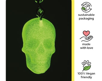 Può includere: Un ciondolo a forma di teschio verde brillante con una superficie strutturata è appeso a una catena di metallo scuro. Il teschio è di un vibrante colore verde neon. L'immagine include anche icone per imballaggi sostenibili, fatto con amore e 100% vegano.