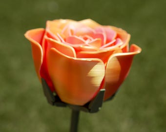 Può includere: Primo piano di una rosa arancione e rosa con uno stelo verde. I petali della rosa sono stratificati e hanno un aspetto strutturato. Lo sfondo è un verde sfocato.