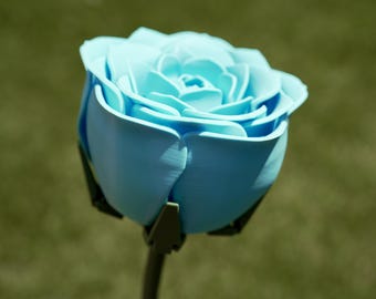 Può includere: Una rosa azzurra con petali dettagliati e uno stelo verde scuro. Il fiore artificiale è in piena fioritura, mostrando il suo design intricato. La rosa è ambientata su uno sfondo verde sfocato, evidenziandone il colore e la forma.