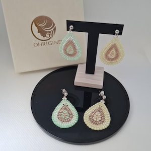 Op de afbeelding: Druppelvormige oorbellen van kralen in mintgroen en crème, gepresenteerd op een zwarte standaard. De oorbellen hebben een zilveren pin en worden gepresenteerd met een merkdoos. Een stijlvol accessoire.