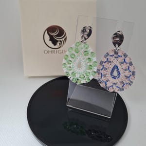 Könnte beinhalten: Tropfenförmige Ohrringe mit grünen und rosa Perlen, präsentiert auf einem transparenten Ständer. Die Ohrringe weisen aufwendige Perlenarbeiten auf und werden mit einer Markenbox präsentiert. Auf einem schwarzen, kreisförmigen Sockel.