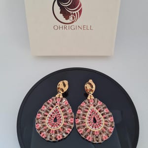 Op de afbeelding: Een paar druppelvormige oorbellen met goudkleurige bovenkanten en een kralenontwerp in roze en bruin. De oorbellen worden gepresenteerd op een zwart oppervlak, met een doos op de achtergrond met het woord "OHRIGINELL".