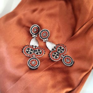 Peut inclure: Une paire de boucles d'oreilles pendantes composées de motifs circulaires ornés de petites perles orange et noires entourant des cristaux gris centraux. Elles incluent une pièce en métal argenté texturé reliant le clou à la partie inférieure.