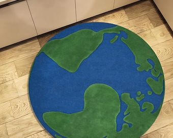 Puede incluir: Una alfombra redonda que representa la Tierra, con océanos azules y continentes verdes. La alfombra está colocada sobre un suelo de madera, con una parte de un mueble blanco visible en el fondo. El diseño de la alfombra es una representación lúdica del planeta.