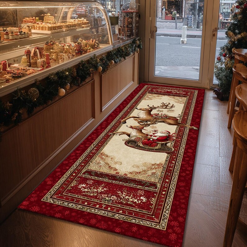 Op de afbeelding: Een rechthoekig kersttapijt met een rode rand en een cr&egrave;mekleurig middenstuk. Het middenstuk toont de kerstman in een slee getrokken door rendieren. Het tapijt ligt voor een deuropening, met een bakkerijvitrine zichtbaar.