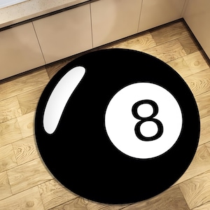 May include: A circular black rug featuring a white number eight and a white curved stripe, similar to a pool ball. The rug is placed on a light brown wooden floor.