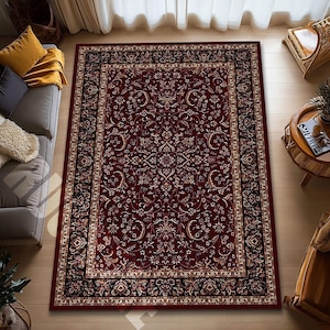 Puede incluir: Una alfombra rectangular con un intrincado diseño floral en tonos burdeos, crema y negro. La alfombra presenta un borde detallado y está colocada sobre un suelo de madera. Un sofá y una mesa auxiliar son visibles en el fondo.