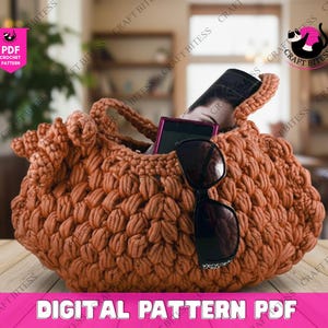 May include: A crocheted brown handbag with a textured, puffy stitch design. The bag contains a smartphone, sunglasses, and a rolled-up item. The words "DIGITAL PATTERN PDF" are displayed in pink text at the bottom of the image.