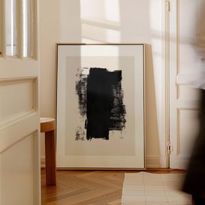 Op de afbeelding: Een ingelijste abstracte kunstprint met een grote zwarte penseelstreek op een beige achtergrond. Het kunstwerk wordt getoond in een kamer met een houten vloer en een gedeeltelijk zichtbare deur en kruk. De lijst is goudkleurig.