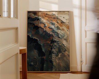 May include: A framed art print depicting a detailed aerial view of a mountainous landscape. The artwork uses a palette of earthy tones, including shades of brown, orange, and grey, with a touch of white. The frame is a light wood colour.