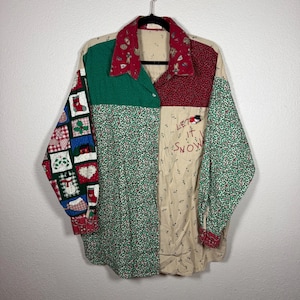 Può includere: Una camicia a maniche lunghe con bottoni e design patchwork con pannelli di tessuto rosso, verde e crema. La camicia presenta motivi a tema natalizio tra cui calze, pupazzi di neve e agrifoglio, con la scritta "LET IT SNOW" ricamata su una tasca.