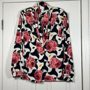 May include: A long-sleeved blouse with a bold floral pattern. The blouse features large pink roses set against a black and white geometric design. The blouse has a draped neckline and buttoned cuffs. The tag reads "Impressions".