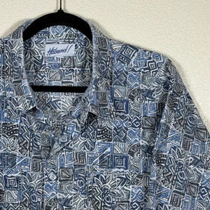 May include: A patterned button-down shirt in shades of blue, grey, and white. The shirt has a geometric design and the brand name "Haband!" on the label. The shirt is hanging on a black hanger.