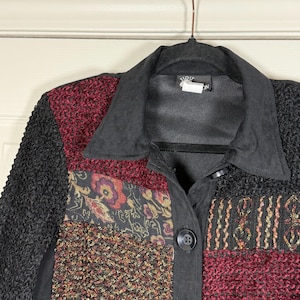 May include: A patchwork jacket with a black collar and button closure. The jacket features panels of textured black fabric, burgundy, and patterned fabric with floral and paisley designs. The jacket is hanging on a brown hanger.