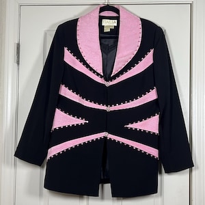 Może przedstawiać: Czarny żakiet z różowymi akcentami i zdobieniami. Żakiet posiada różowy kołnierz i dekoracyjne różowe paski z przodu, ozdobione małymi, błyszczącymi detalami. Etykieta pokazuje "ITALIA".