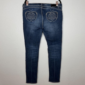 May include: Dark blue denim jeans featuring a faded wash and a straight-leg cut. The back pockets are shaped like apples and are decorated with intricate silver-toned embroidery and small metallic studs, creating a detailed pattern on the fabric.