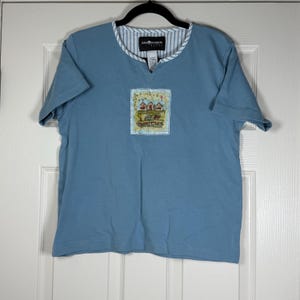 Peut inclure: T-shirt bleu clair à manches courtes avec un col en V et un petit patch brodé. Le patch représente une scène rurale avec des granges rouges et un champ jaune. L'encolure est ornée d'un motif rayé. L'étiquette de la marque est visible.
