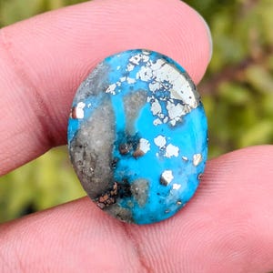 Op de afbeelding: Ovale edelsteen met een levendige turquoise basis, bespikkeld met zilveren en donkergrijze insluitsels. Het gepolijste oppervlak reflecteert het licht en benadrukt de natuurlijke patronen. Een unieke, opvallende steen voor sieraden of verzamelingen.