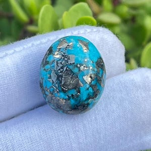 Può includere: Una pietra preziosa turchese di forma ovale caratterizzata da un vibrante colore di base azzurro cielo con prominenti inclusioni metalliche irregolari di pirite color argento sparse sulla superficie, creando un netto contrasto cromatico.