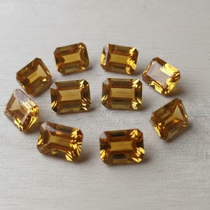Può includere: Una collezione di dieci gemme rettangolari sfaccettate. Le gemme sono di un caldo colore giallo dorato e disposte in un motivo circolare. Le gemme hanno una qualità brillante e riflettente, suggerendo che siano tagliate per massimizzare la rifrazione della luce.
