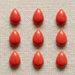 Può includere: Nove cabochon lisci a forma di goccia in una vibrante tonalità arancione-rossa. Ogni pezzo presenta una superficie lucida e arrotondata con un riflesso brillante, disposti in una griglia uniforme tre per tre su un tessuto testurizzato.