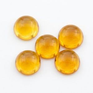 Könnte beinhalten: Fünf ovale, bernsteinfarbene Edelsteine liegen auf weißem Grund. Die polierten Cabochons reflektieren das Licht und zeigen ihre glatten Oberflächen. Geeignet für Schmuckherstellung oder andere Bastelprojekte.
