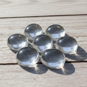 May include: Seven clear, rounded glass pebbles are arranged on a light-coloured wooden surface. The pebbles have a smooth, polished appearance, reflecting light. The overall effect is simple and elegant.