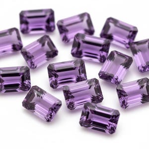 May include: A selection of rectangular-cut amethyst gemstones. The gems are a deep purple colour and have a polished, reflective surface. The stones are scattered across a white background, showcasing their clarity and colour.