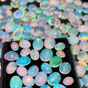 May include: A collection of oval-shaped opals in various sizes, displaying a range of colors including blue, green, pink, and white. The opals are set against a black background, showcasing their iridescent qualities. The gemstones are arranged in a tray.