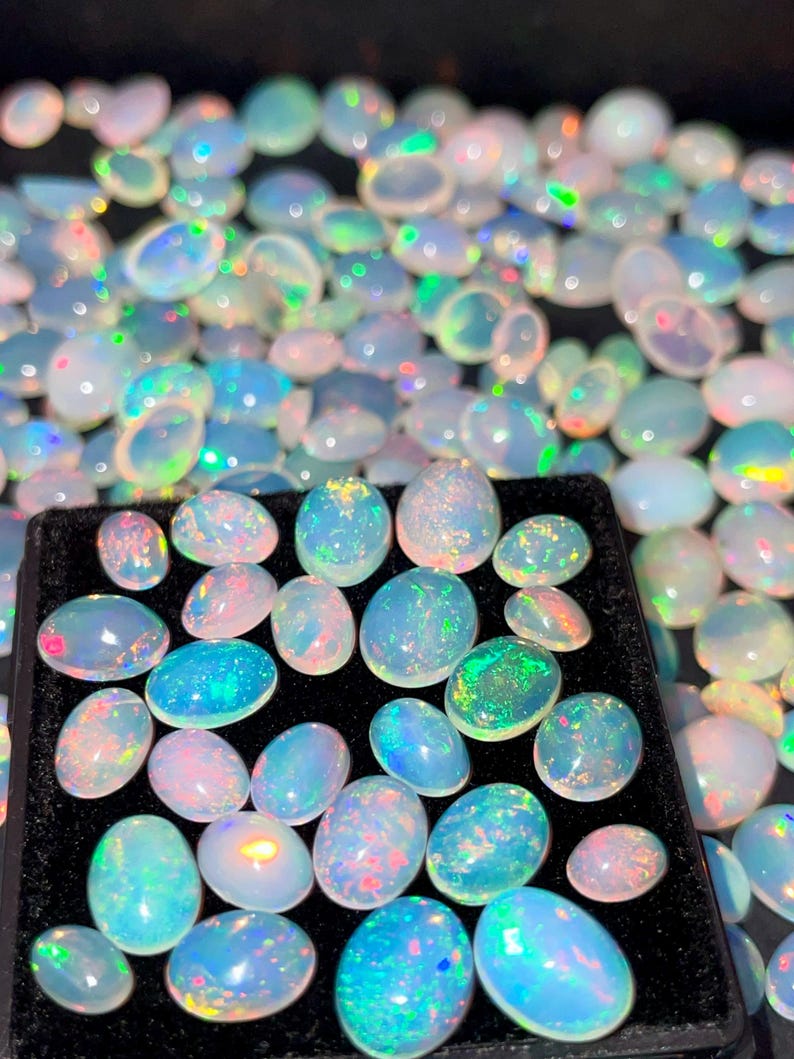 May include: A collection of oval-shaped opals in various sizes, displaying a range of colors including blue, green, pink, and white. The opals are set against a black background, showcasing their iridescent qualities. The gemstones are arranged in a tray.