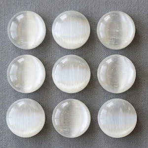 Peut inclure: Neuf cabochons circulaires blanc translucide disposés en une grille de trois par trois. Chaque pièce présente une surface lisse et bombée avec un effet chatoyant qui reflète la lumière en une bande verticale au centre des pierres.