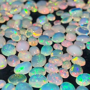 Könnte beinhalten: Eine Sammlung ovaler Opale in verschiedenen Größen. Die Opale zeigen eine Reihe von Farben, darunter Weiß, Blau, Grün und Rosa, mit schillernden Blitzen. Die Edelsteine sind auf einer dunklen Oberfläche verstreut.